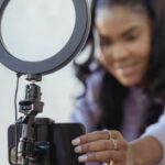 Style Blog - Cheerful young African American female blogger in stylish sweater smiling while setting up camera of smartphone attached to tripod with ring light before recording vlog