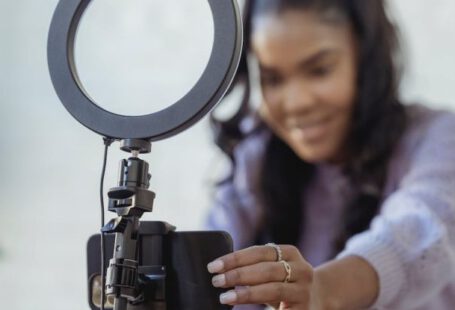 Style Blog - Cheerful young African American female blogger in stylish sweater smiling while setting up camera of smartphone attached to tripod with ring light before recording vlog