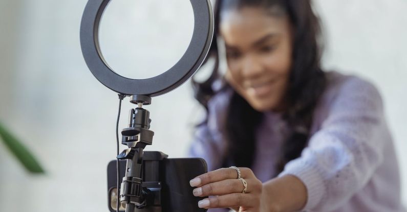 Style Blog - Cheerful young African American female blogger in stylish sweater smiling while setting up camera of smartphone attached to tripod with ring light before recording vlog