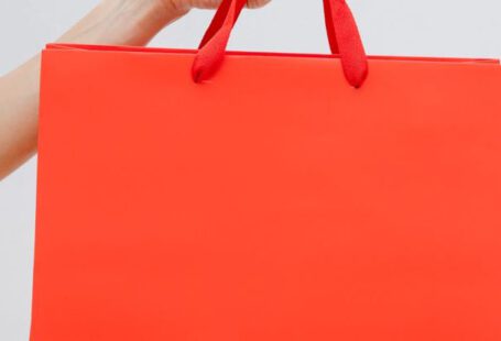 Clearance Sales - Crop unrecognizable female showing red paper shopping bag in hand against white wall