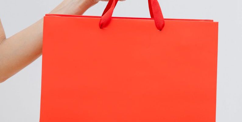 Clearance Sales - Crop unrecognizable female showing red paper shopping bag in hand against white wall